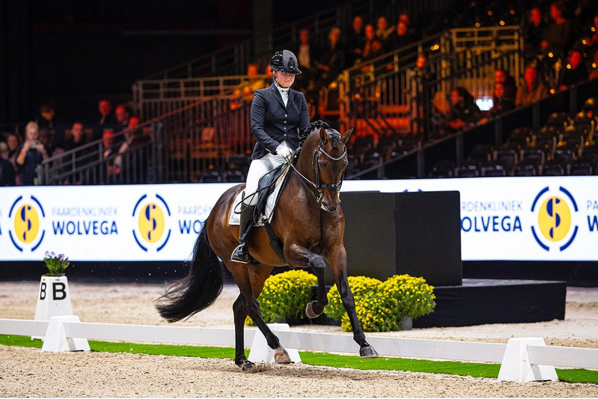 Fleur Prinsen riding an extended canter with her bay colored horse in an indoor arena, wearing a JuulC show jacket